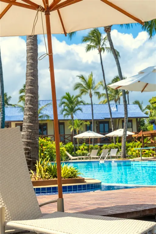 Prédio de telhado azul entre coqueiros, com piscina e guarda-sóis à frente
