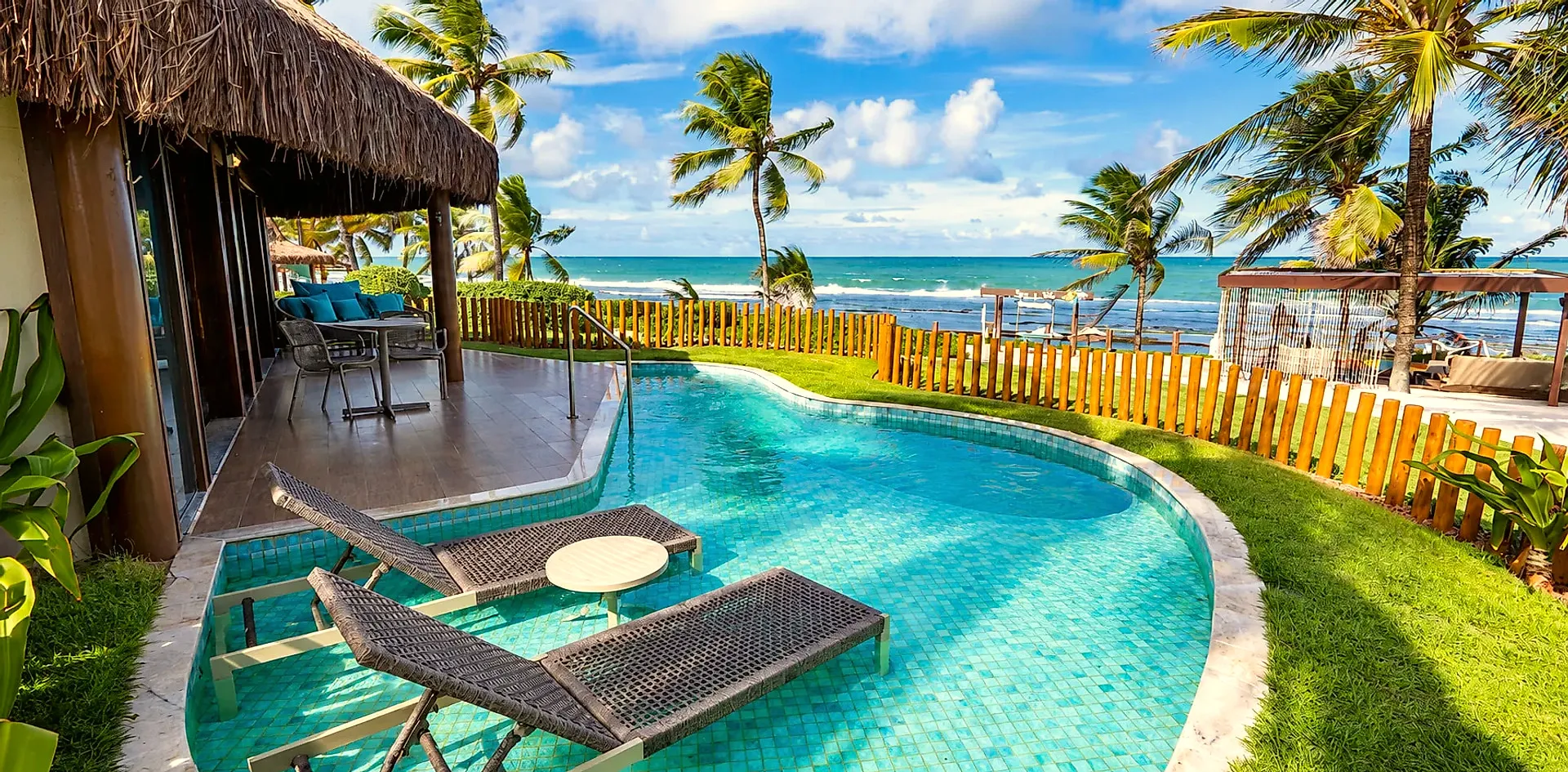 Piscina privativa do Bangalô Exclusive com duas espreguiçadeiras na água, varanda com telhado de palha, gramado e o mar ao fundo