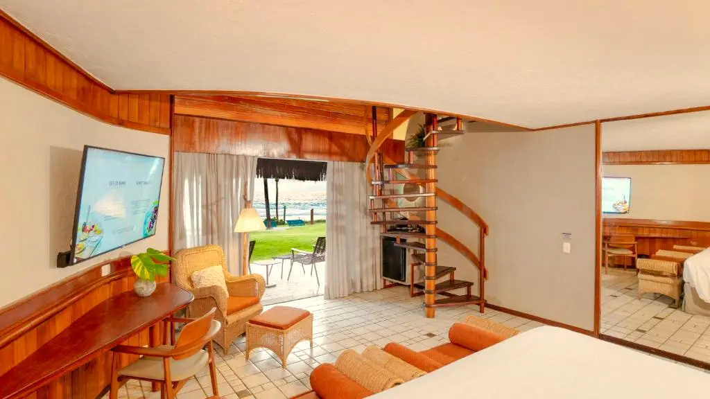 Sala do bangalô com vista para o mar pela porta de vidro e escada para o mezanino