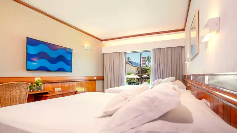 Cama com travesseiros e TV na parede do Apartamento Classic, com janela para a piscina