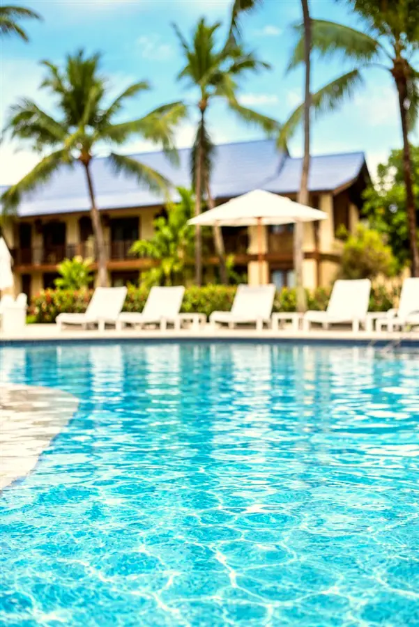 Espreguiçadeiras brancas à beira da piscina, com guarda-sol e prédios ao fundo
