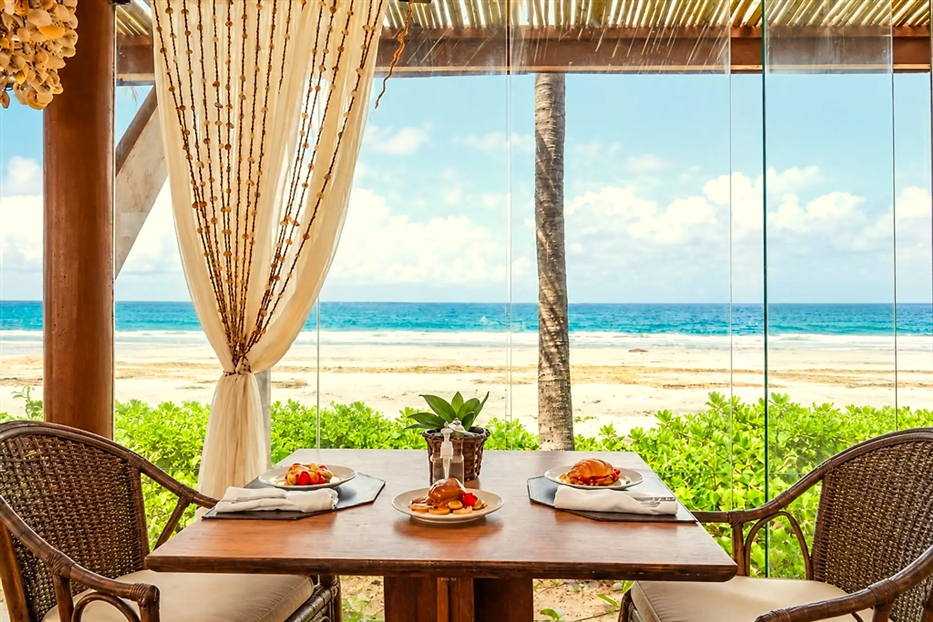 Mesa posta de frente para a praia, com cortinas e a areia ao fundo