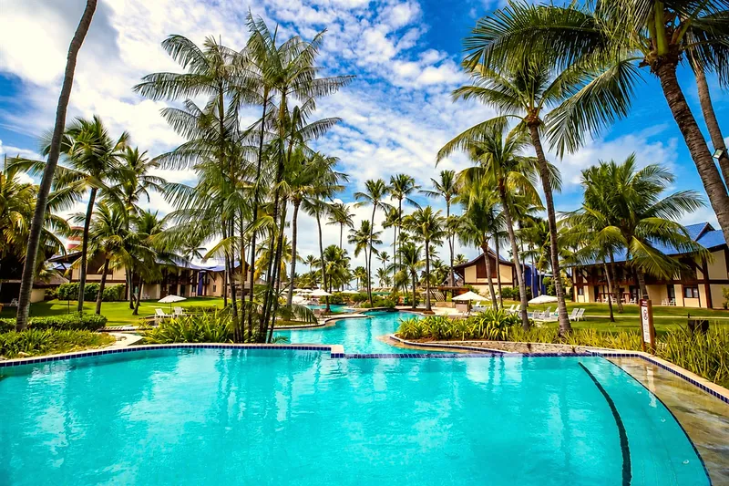 Piscina de água azul-turquesa cercada de coqueiros, jardins e guarda-sóis
