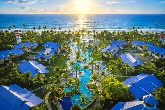 Vista aérea do resort ao amanhecer, com bangalôs de telhado azul, piscinas entre coqueiros e o mar ao fundo